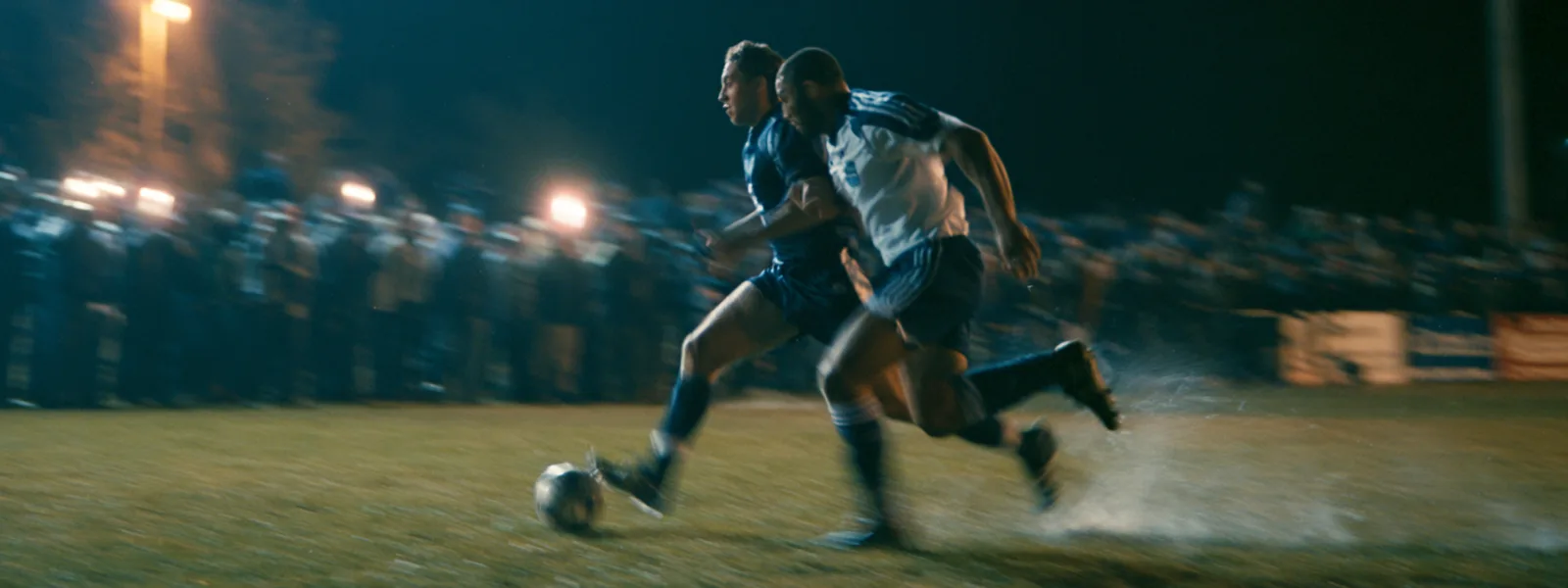 Twee voetballers die zij aan zij naar de bal sprinten, verlichte tribunes bij avond